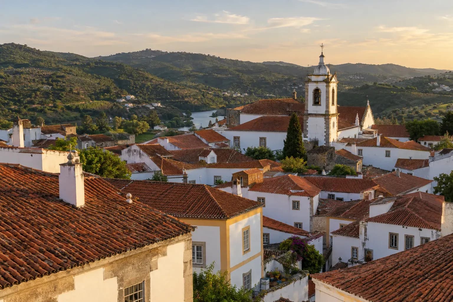 Telhados de barro e fachadas brancas de uma vila portuguesa, com uma torre de igreja e colinas ao fundo.