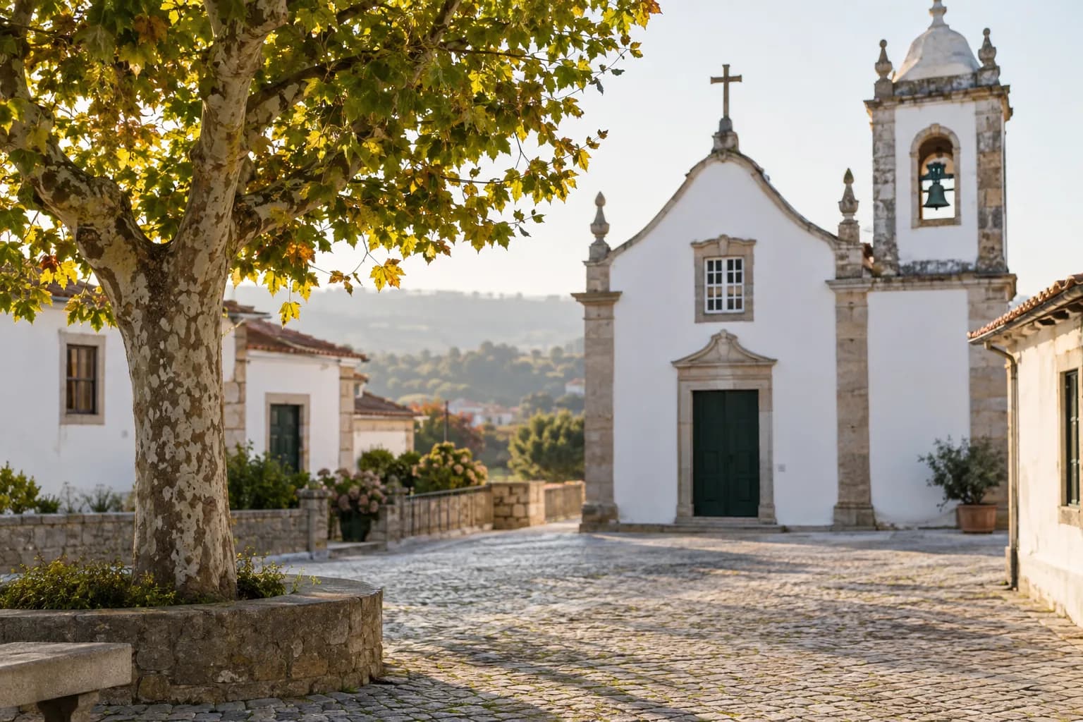 Praça calcetada numa aldeia portuguesa, com uma árvore de copa grande, uma igreja de fachada branca e torre sineira.