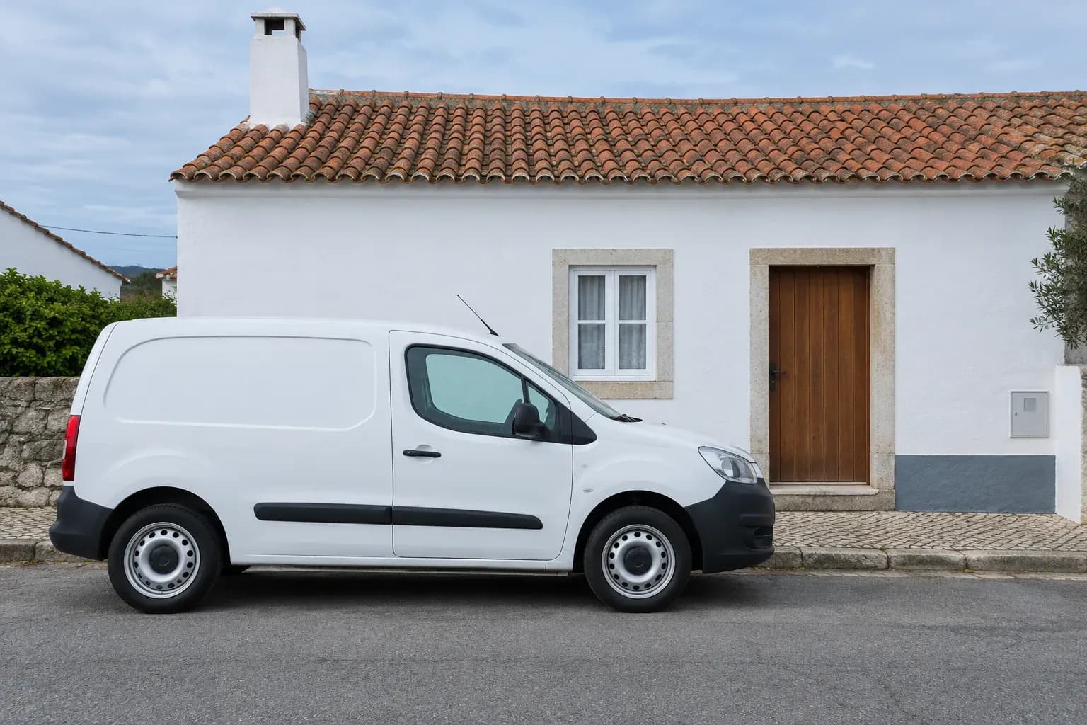 Carrinha branca de serviço estacionada em frente de uma moradia caiada, com telhado de telha e porta de madeira.